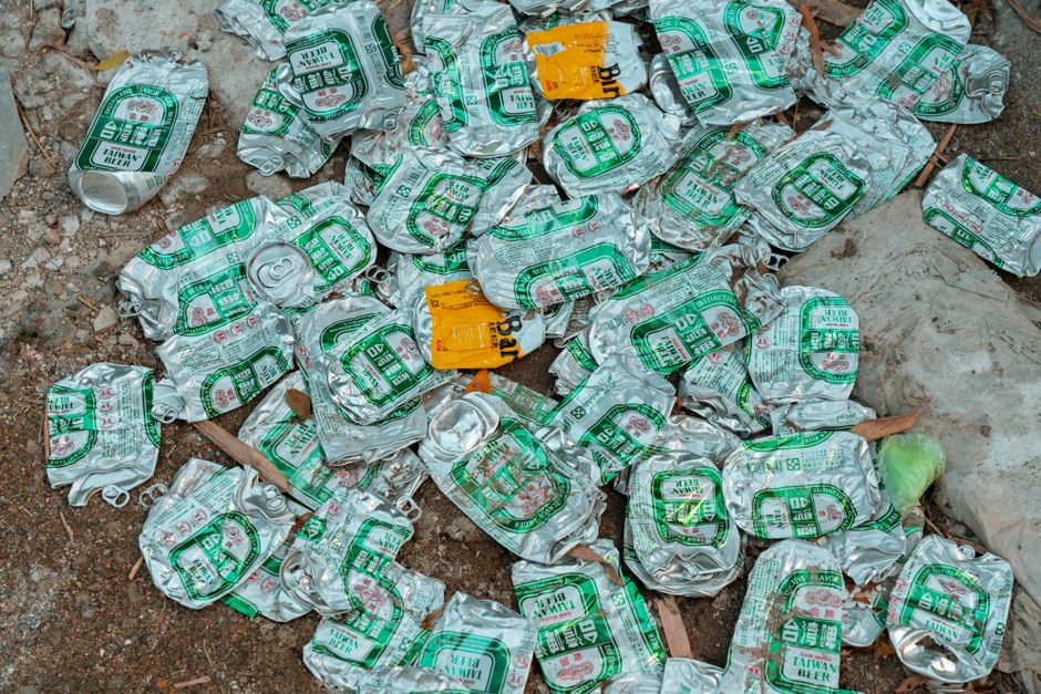 A pile of crushed aluminium cans, predominantly silver in color with green printed labels, is scattered on a rough outdoor ground surface composed of soil, small rocks, and scattered debris. The cans,